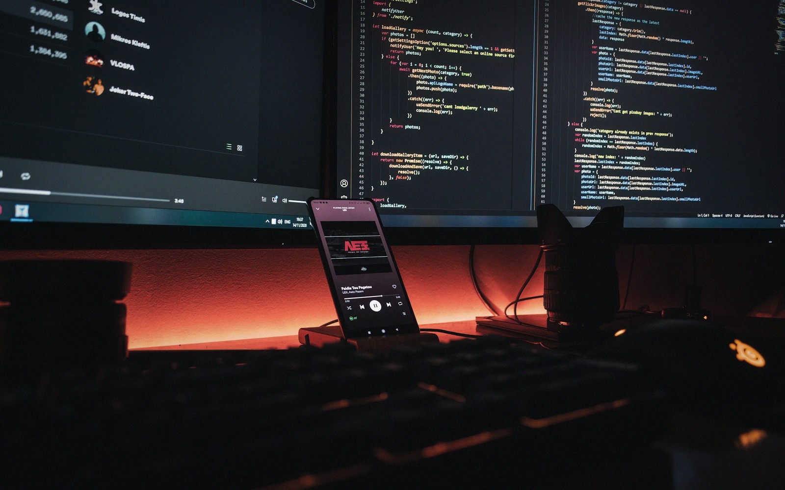 Smartphone playing music on a desk beside monitors displaying lines of code